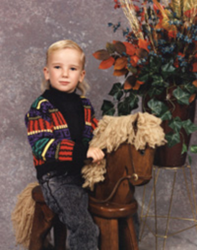 A young boy sitting on a toy horse with a plant backdrop.