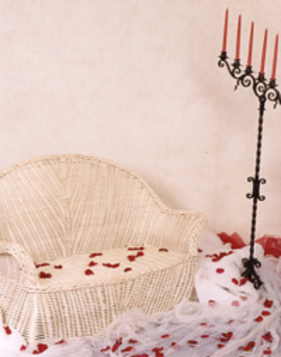 Cozy wicker loveseat with red rose petals scattered on and around it against a soft pink wall.