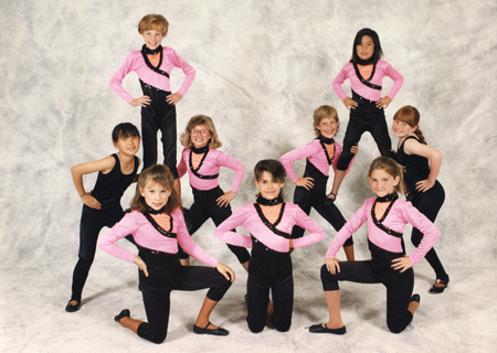 A group of nine dancers in coordinated pink and black outfits striking dynamic poses.