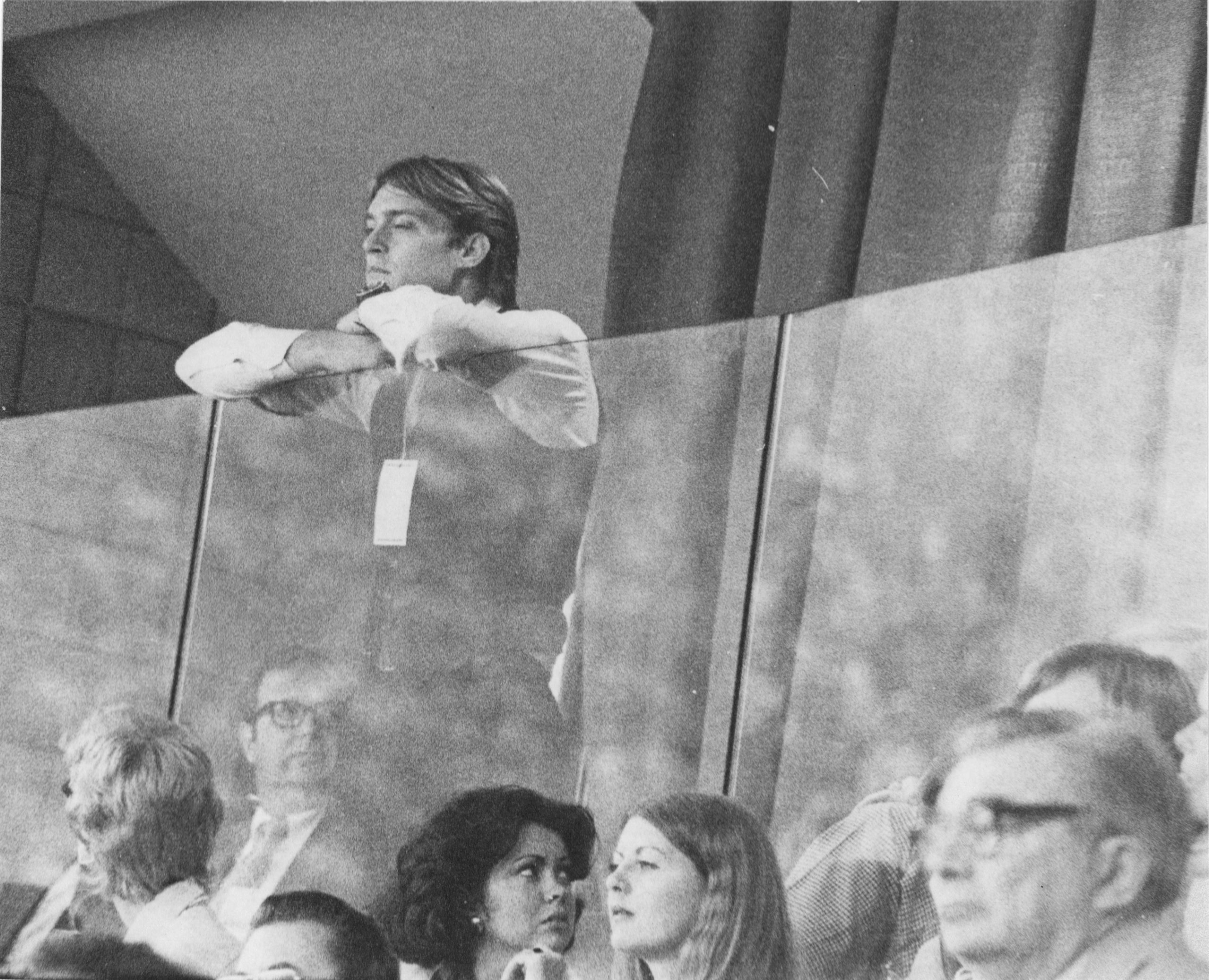 A person speaking in front of an audience with a microphone and glass barrier.