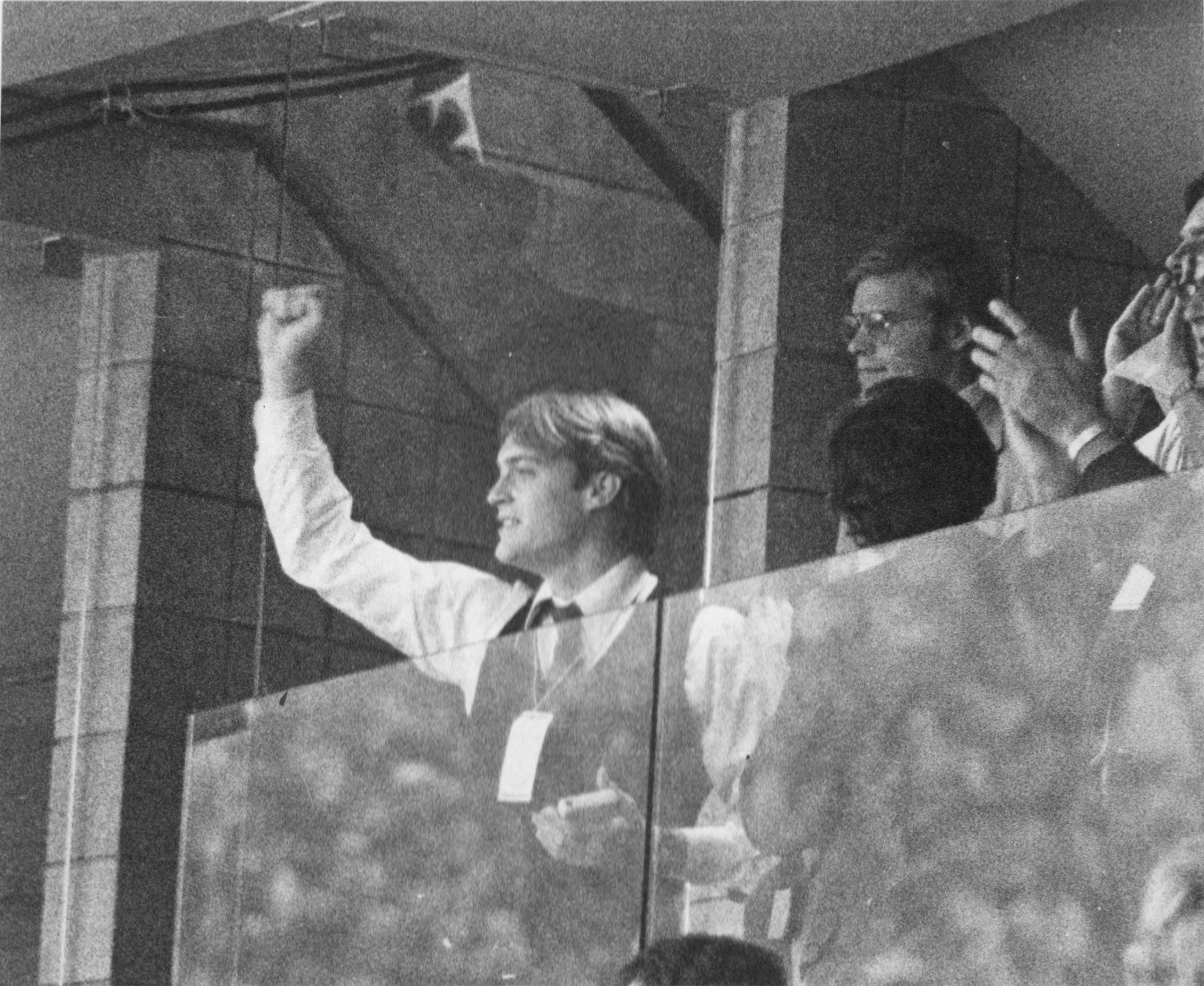 Two men enthusiastically waving from a balcony or enclosed space.