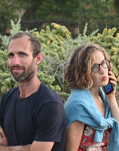 Two people standing back-to-back outdoors, one talking on a phone.