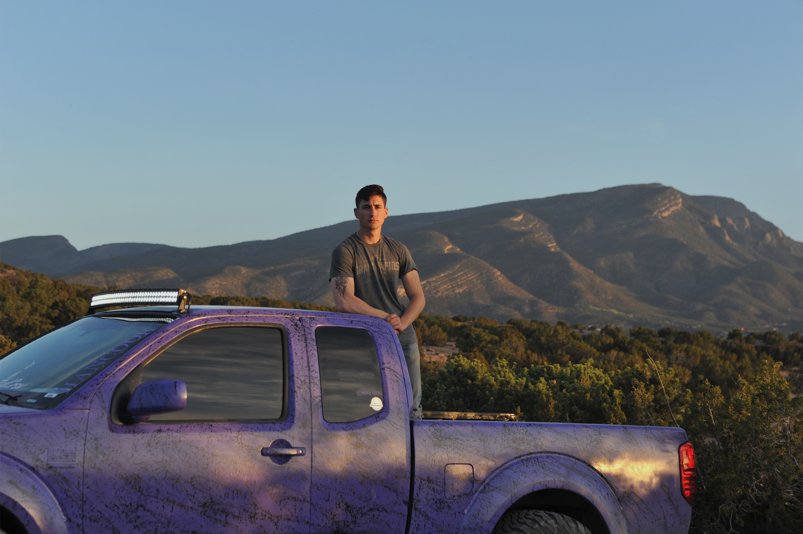 A man sitting on the roof of a purple pickup truck with mountains in the background.