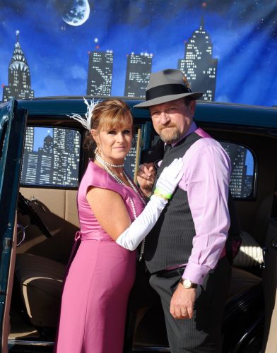 A couple dressed in vintage attire posing by a classic car in a city.